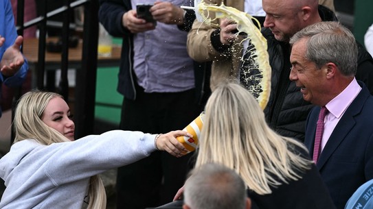 A young woman throws a milkshake at the face of a politician in a crowd.