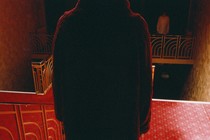 Filling the middle of the frame is the back of a person standing on a staircase, facing another person across the hall. The carpet is red, and shadows are deep.