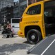 A woman in a wheelchair rolls past a taxi with an open ramp.