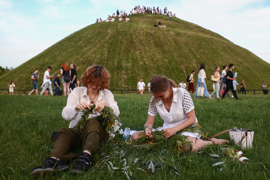 People sit in grass beside a large grass-covered mound, weaving wreaths made of flowers.