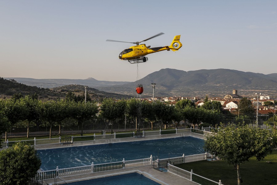 A firefighting helicopter collects water from a swimming pool with a large bucket that hangs from a cable.