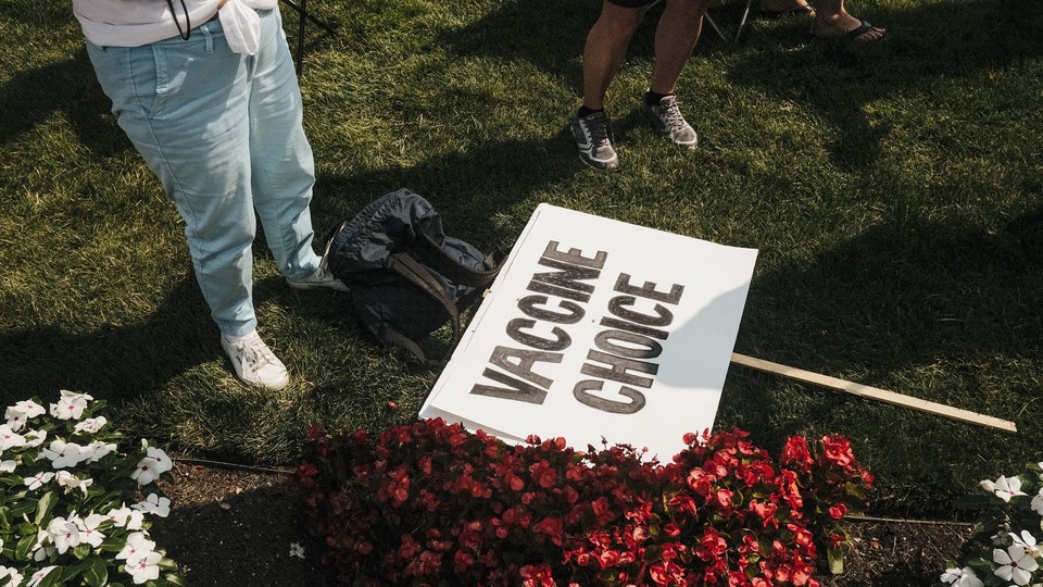 A photo of the legs of a person standing on a patch of grass next to a placard on the ground that reads "Vaccine Choice"