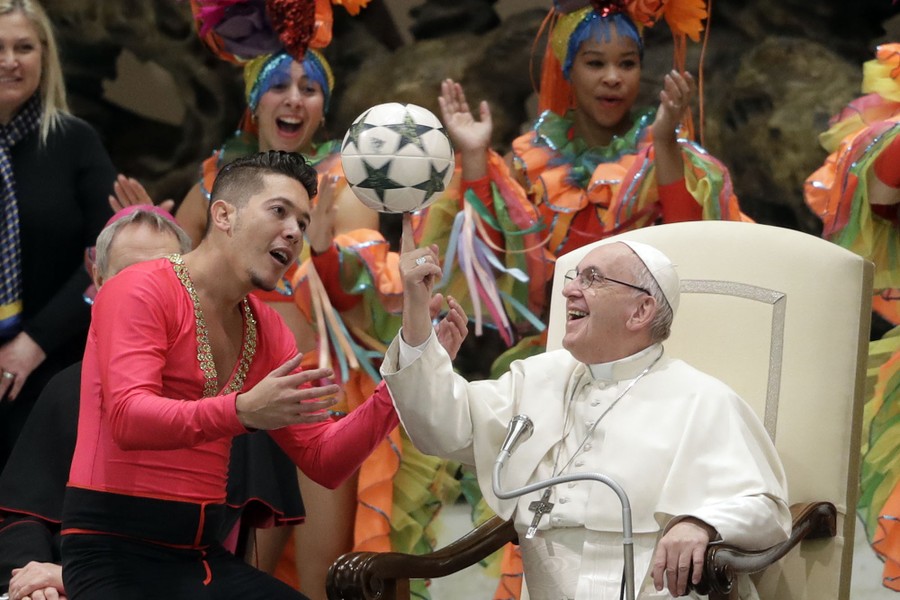 Pope Francis, seated in a large chair and surrounded by performers, twirls a soccer ball on a finger, helped by another performer.