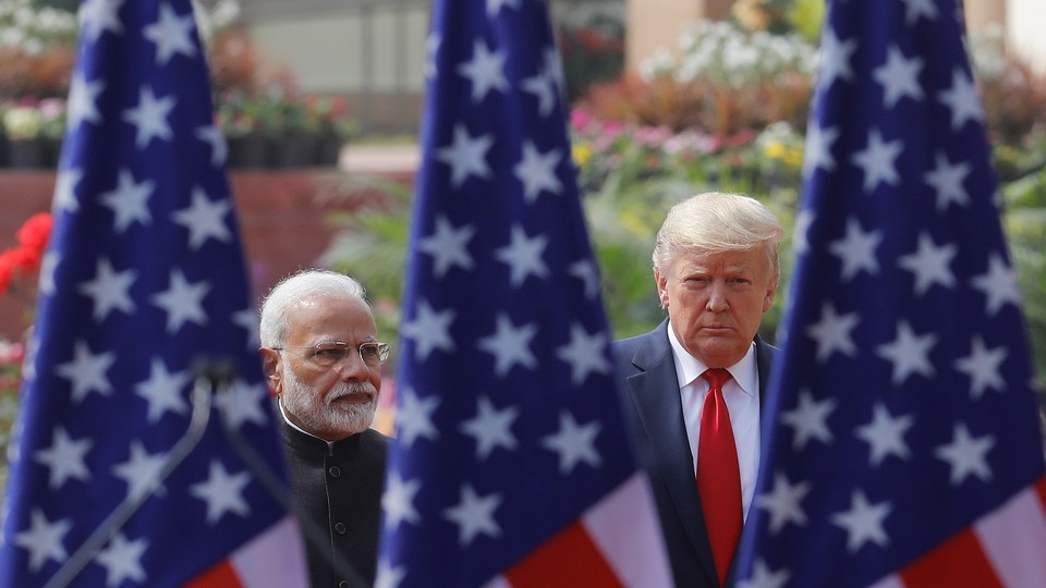 Color photo of Narendra Modi standing next to Donald Trump with three American flags in the foreground