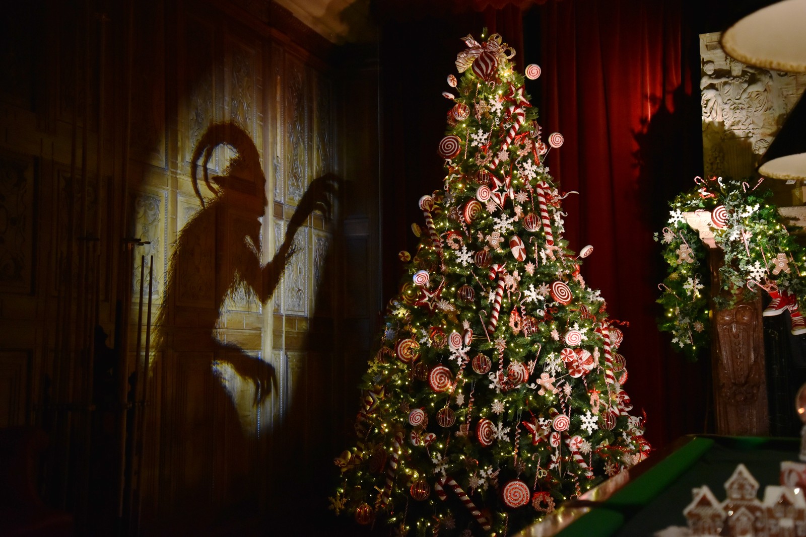 A Christmas display inside a large manor, including a decorated Christmas tree and the shadowy outline of a Krampus figure projected against a wall
