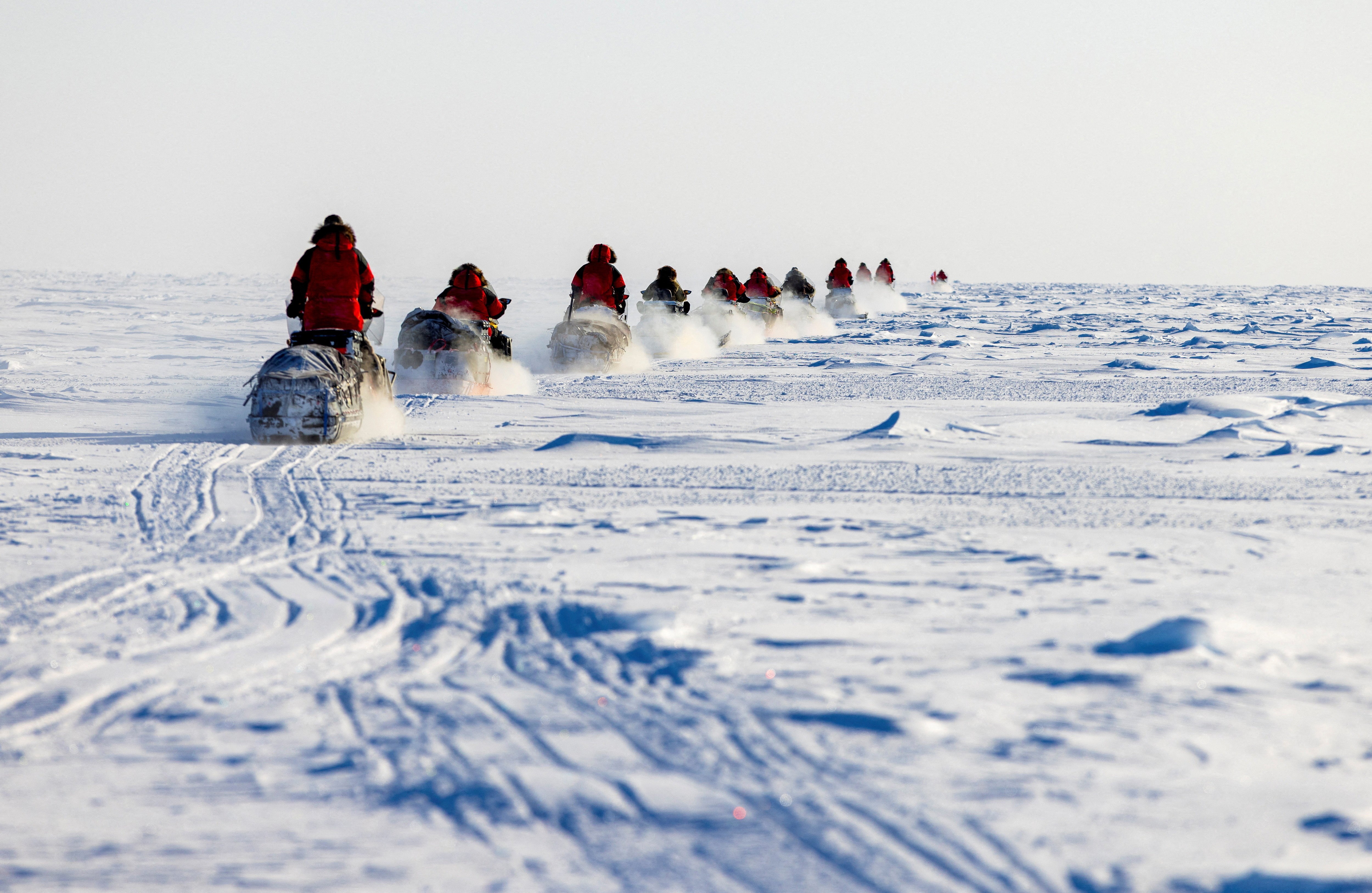 About a dozen people in red parkas ride snowmobiles in a line across a broad, flat expanse of snow and ice.