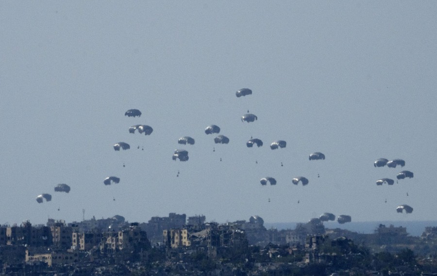 Two dozen parachutes carrying small crates fall toward buildings.
