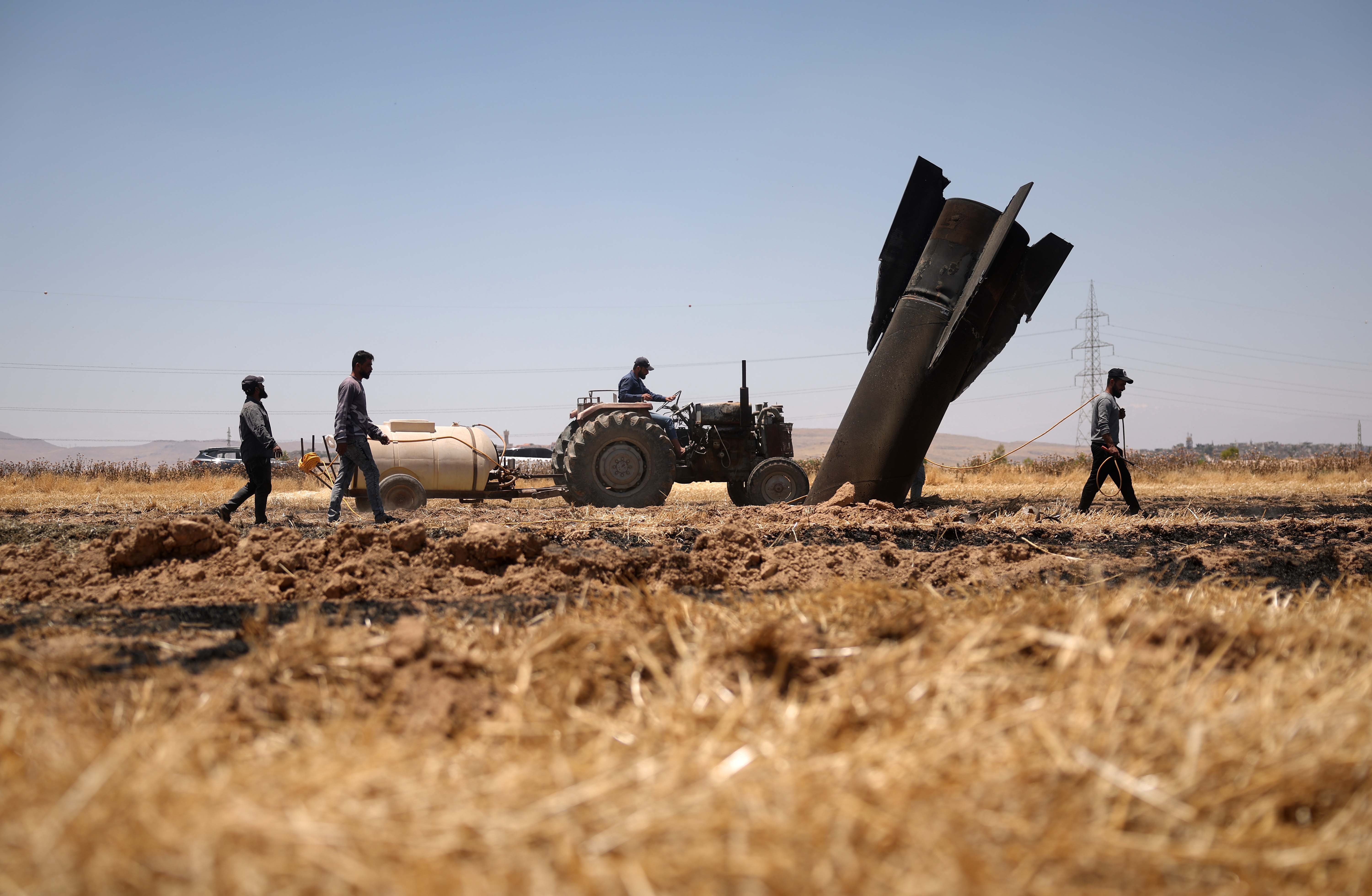 Farmers spray water in a burned agricultural field next to a fallen missile that sticks out of the ground, fins up.