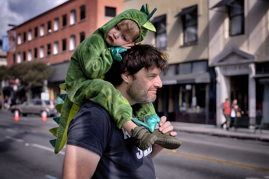 A child in a dinosaur costume rides on his father's shoulders on a street after a parade.