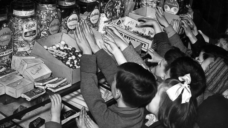 A black and white photograph of a gaggle of children reaching up onto a counter filled with boxes and jars of candy
