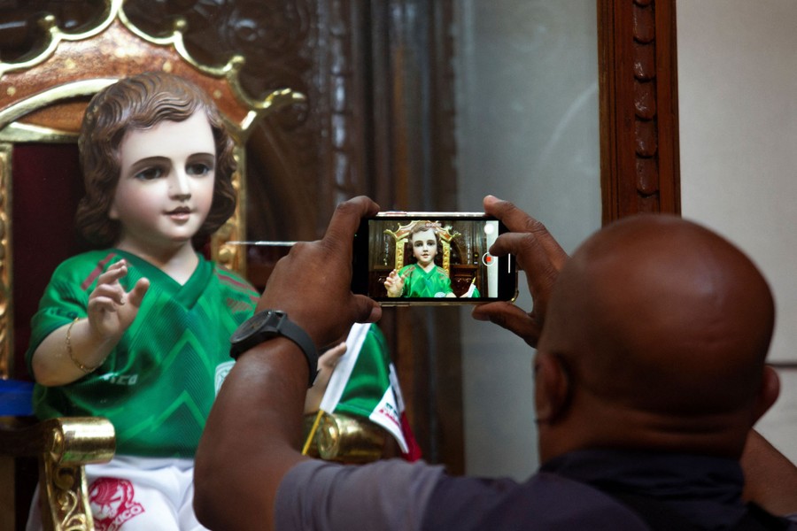 A man takes a picture of a doll dressed in the Mexican national soccer team uniform.
