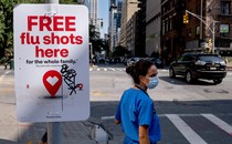 Photo of a woman on a city sidewalk wearing medical scrubs and a mask, next to a sign promoting free flu shots