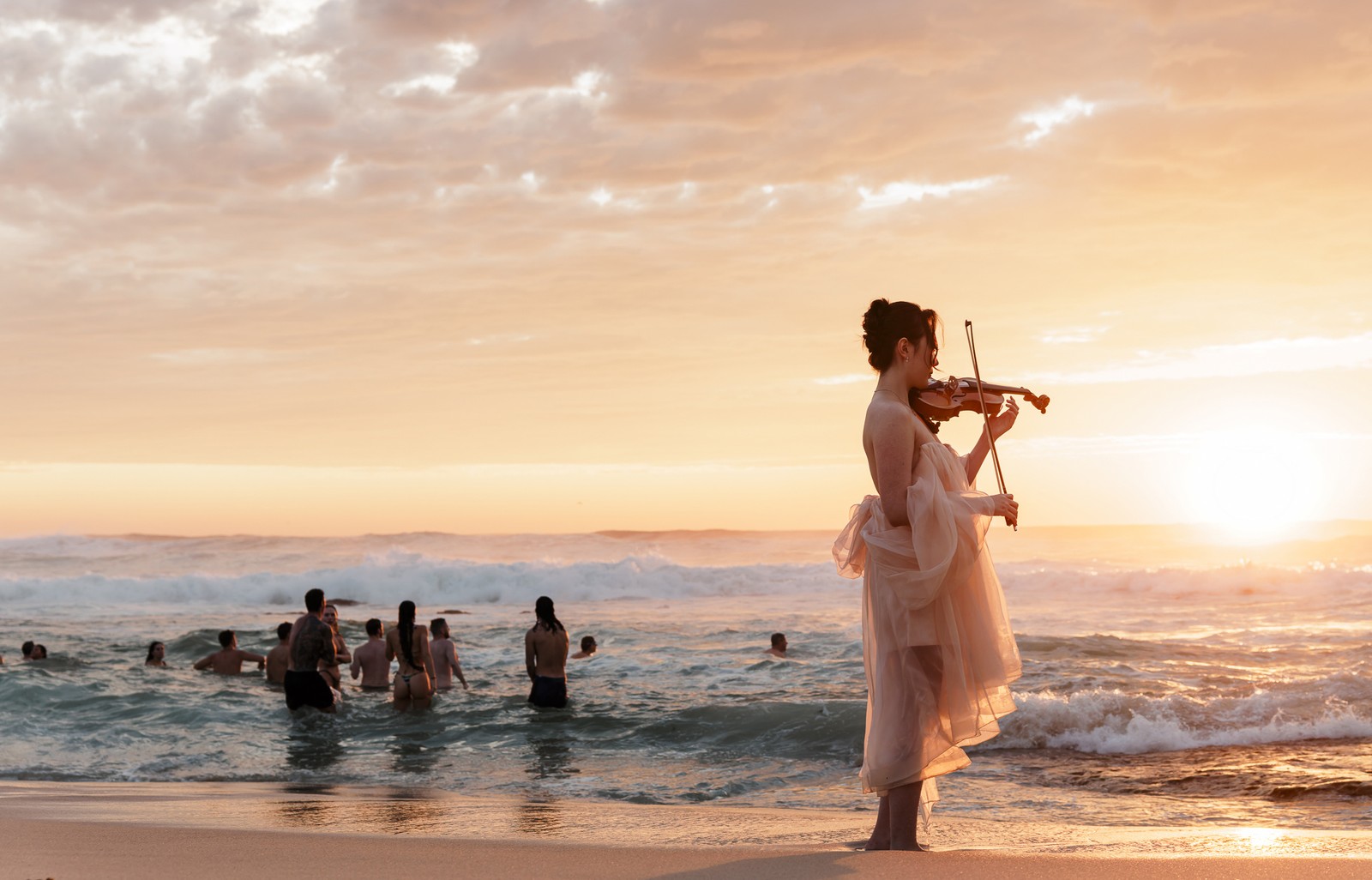 A woman with a violin stands along the surf on a beach, as others play in the waves nearby.