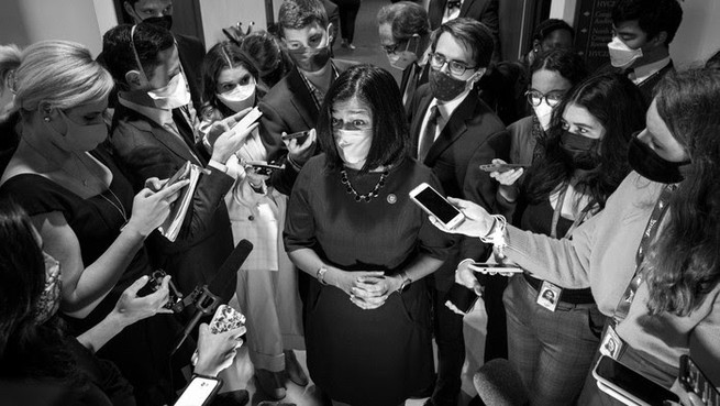 A black and white photo of a masked woman wearing black. She is surrounded by masked reporters holding phones and notebooks.