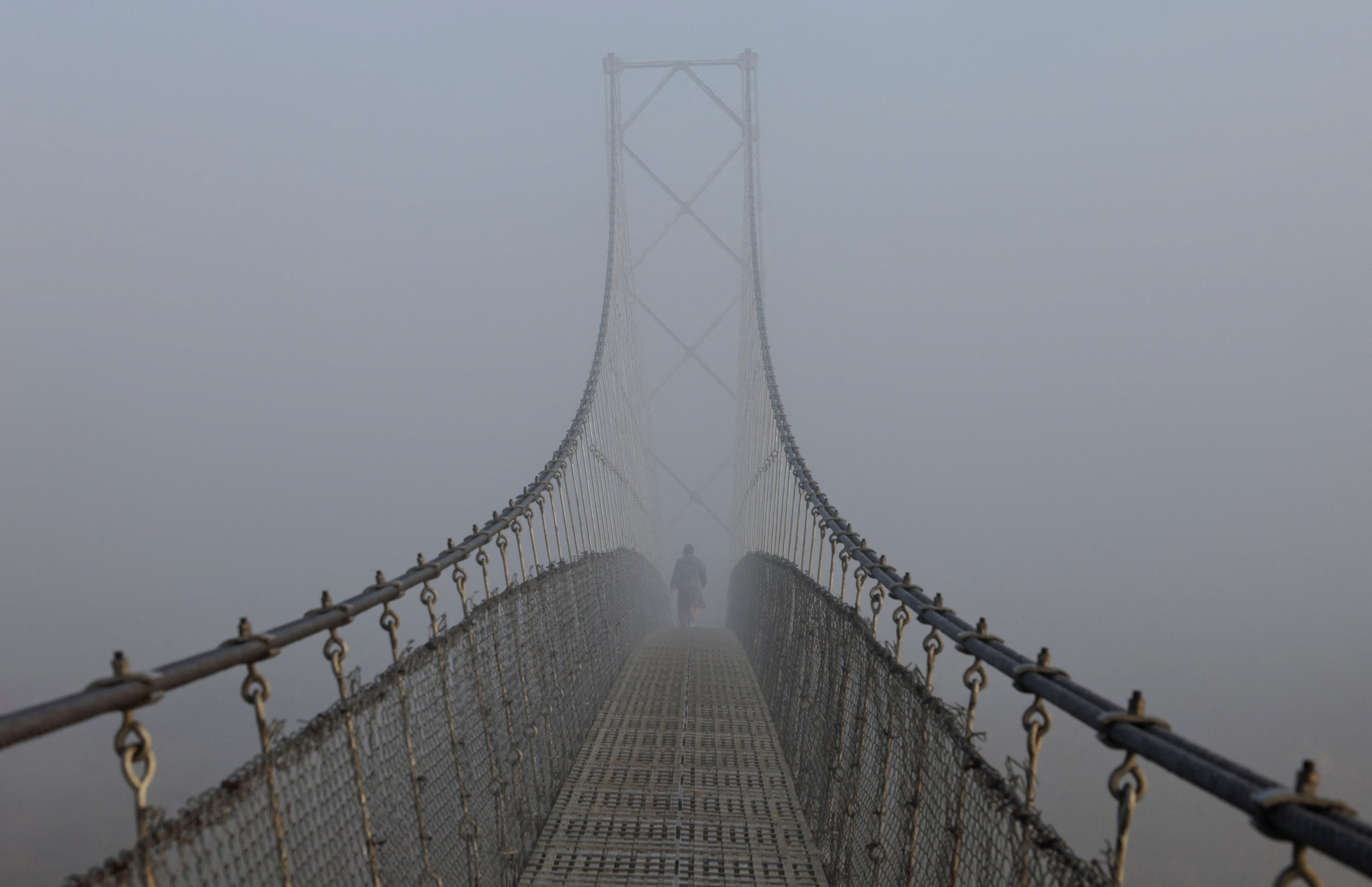 A woman walks on a suspension bridge during a foggy morning.