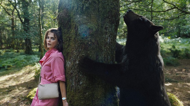 A film still of Cocaine Bear standing on one side of a tree, and a woman hiding on the other