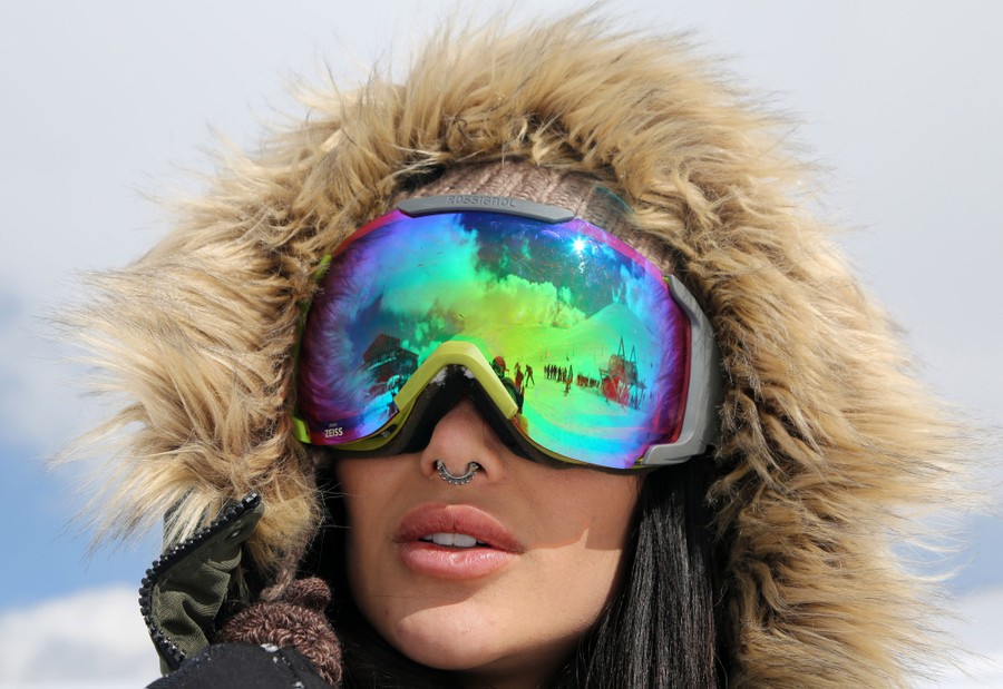 A person wearing a fuzzy hood and large ski goggles stands atop a ski run.