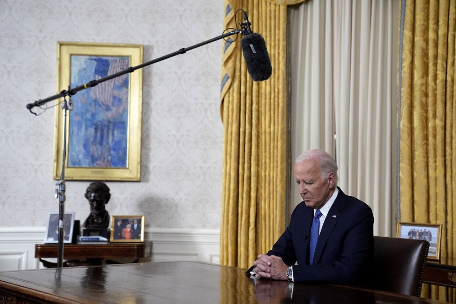 President Joe Biden sits at a desk, head slightly bowed, beneath a boom microphone.
