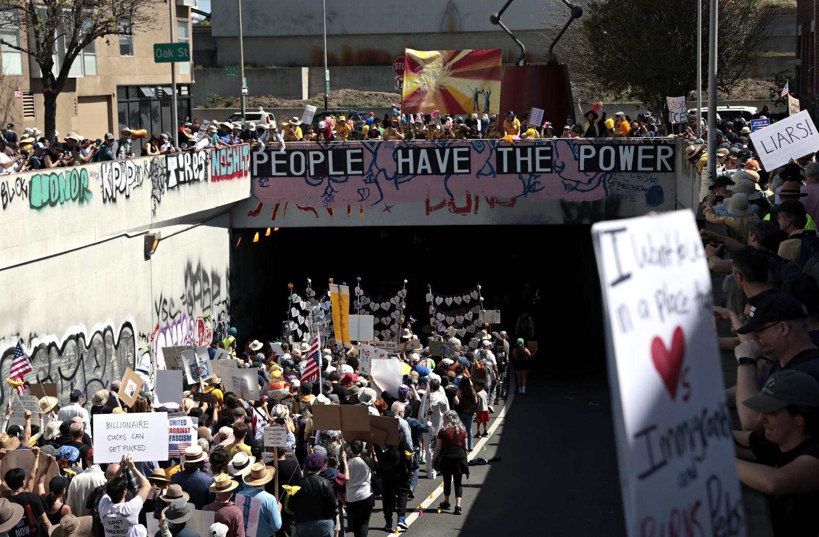 A crowd of protesters, carrying signs, marches on a street beneath an overpass.