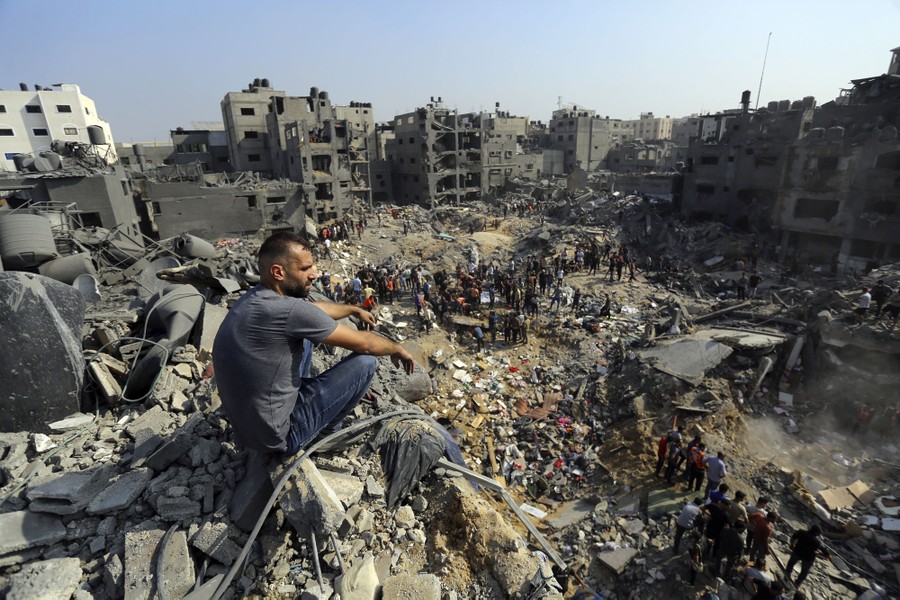 Crowds of people gather among building debris and craters in a bombed-out area in Gaza.