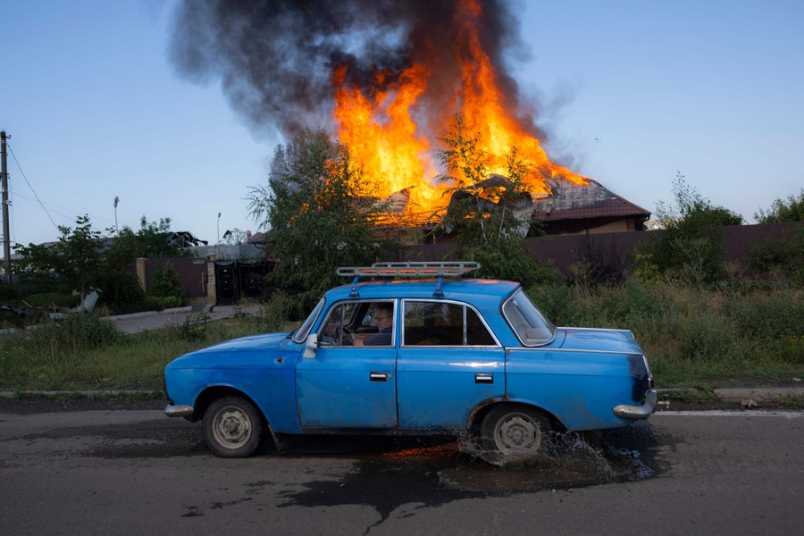 An old blue car drives down a road past a house that has flames erupting from its roof.