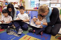 Preschool children sit and read with adults on a colorful rug. 