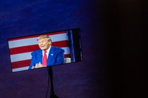 A screen shows Donald Trump speaking in front of an American flag