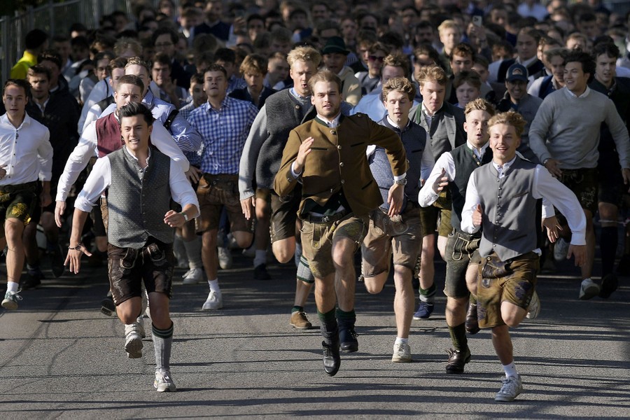 A crowd of mostly young men dressed in lederhosen run at a festival entrance.