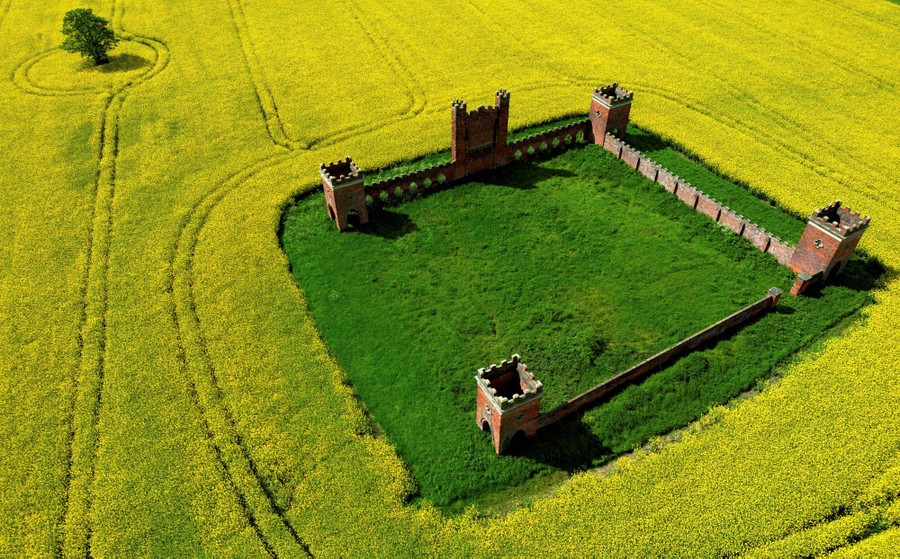 An empty structure made of three crenelated walls and short towers stands on a green patch in a field of yellow plants.
