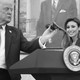 A black-and-white photo of Donald Trump touching a podium microphone while Alina Habba smiles in the background.