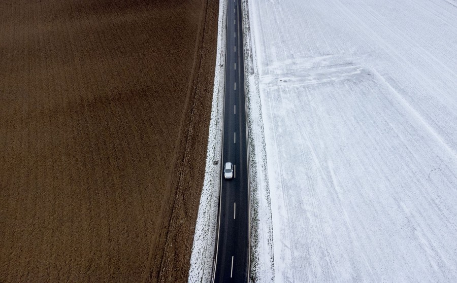 A car passes on a road between a bare field and a snow-covered one.