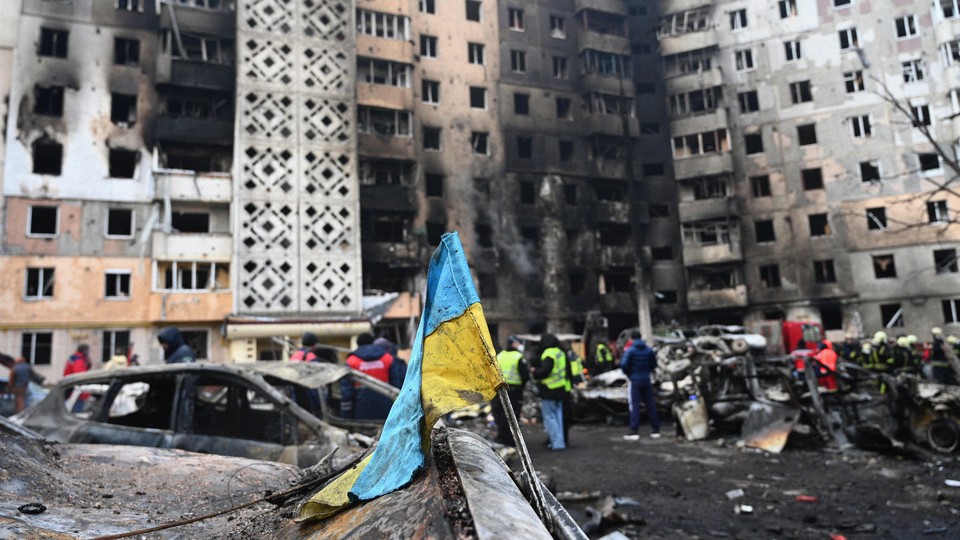 A photo shows bomb-damaged city buildings with emergency workers, burned cars, and a muddy Ukrainian flag in the foreground.