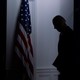 A dark photo of the silhouette of Joe Biden standing by an American flag