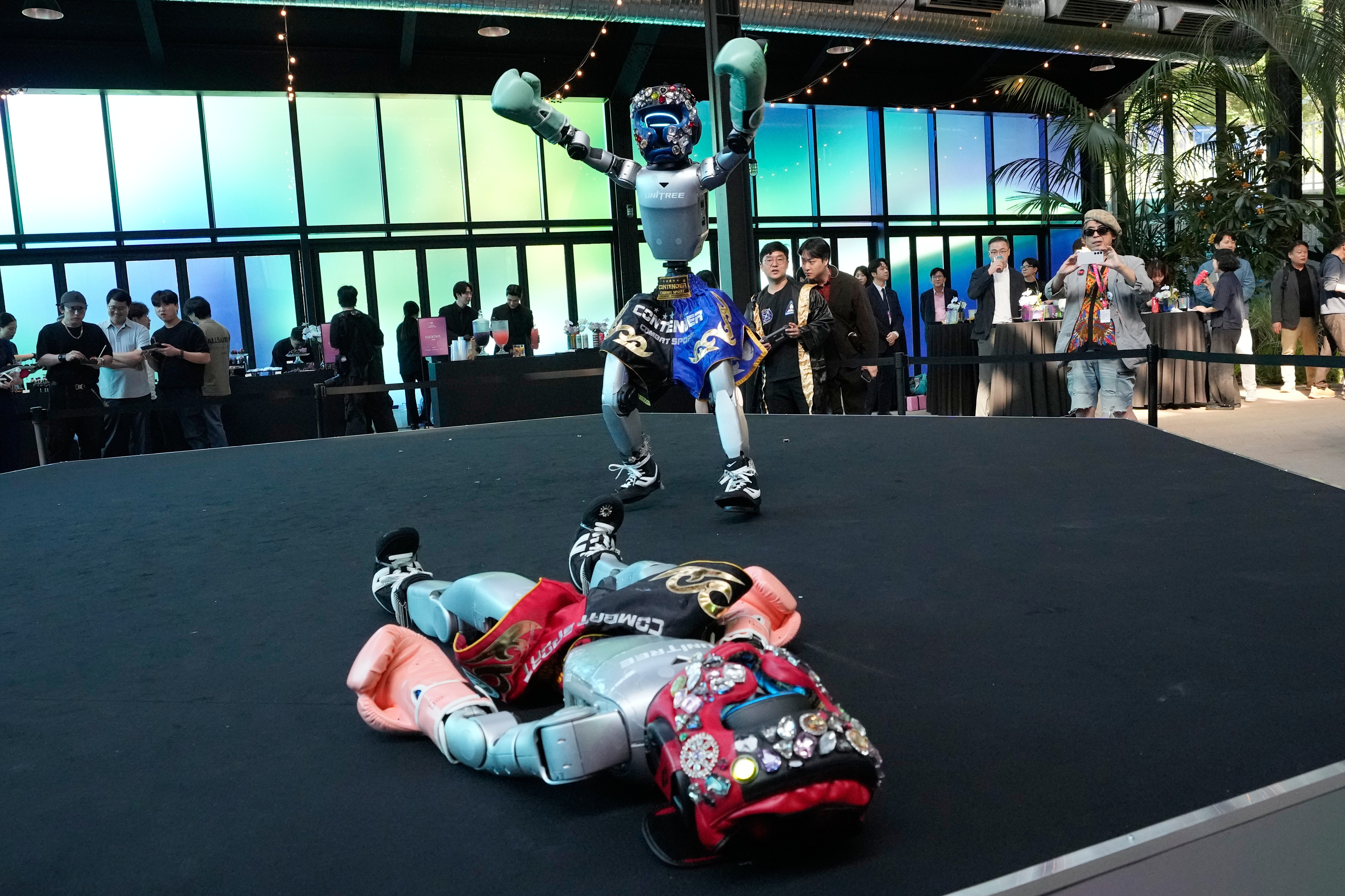 Onlookers watch as one humanoid robot appears to celebrate after knocking down another robot in a boxing match.
