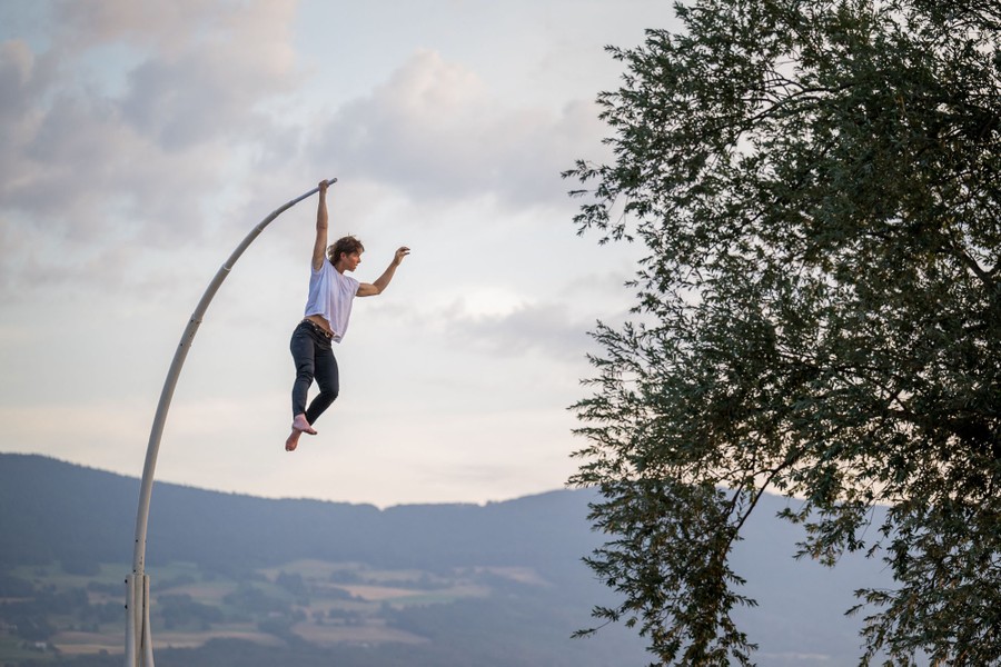 A performer hangs from the end of a tall curved pole, by one hand.