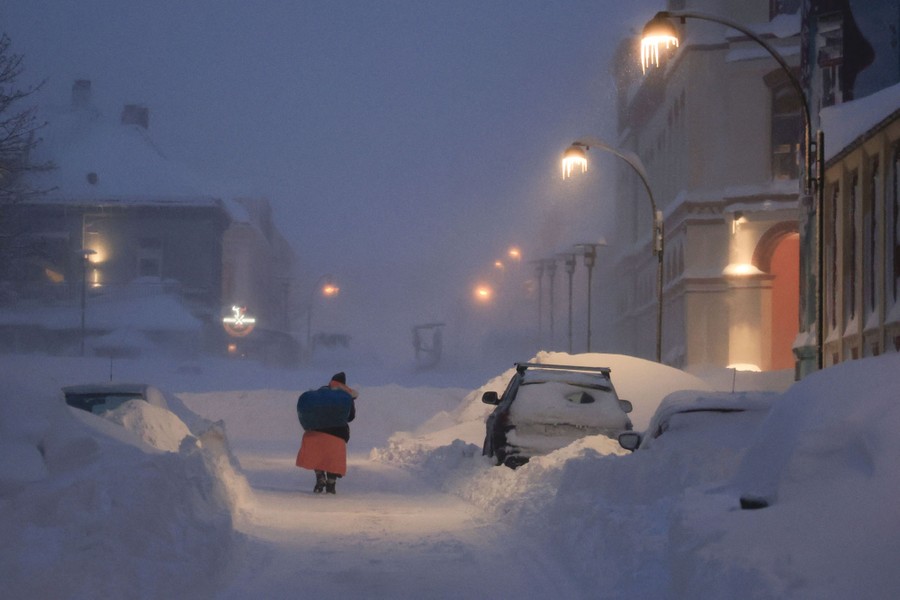 A person walks on a plowed road amid piles of snow and snow-covered buildings.