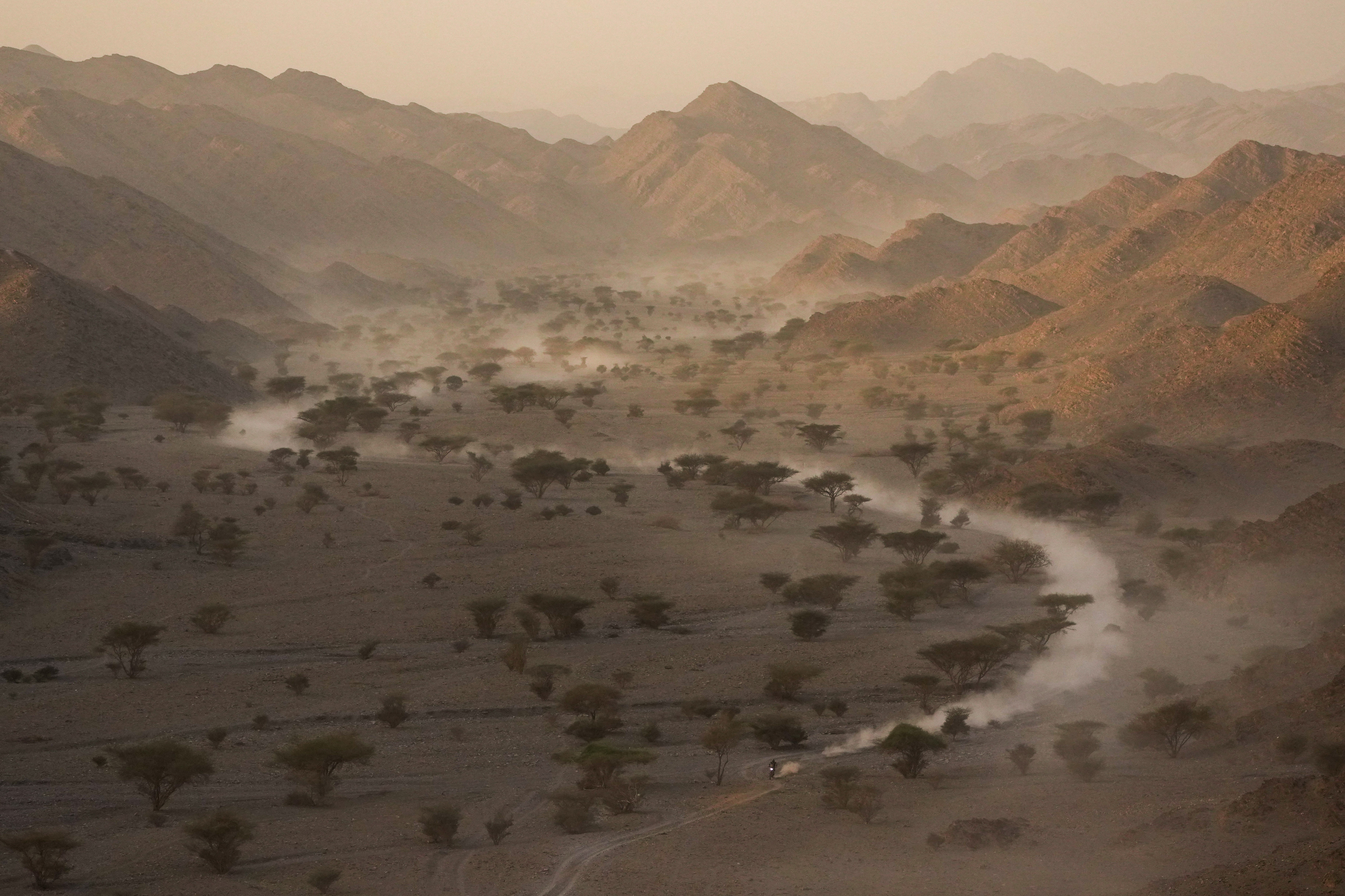 A motorcycle racer rides on a dusty path through a shallow desert valley.