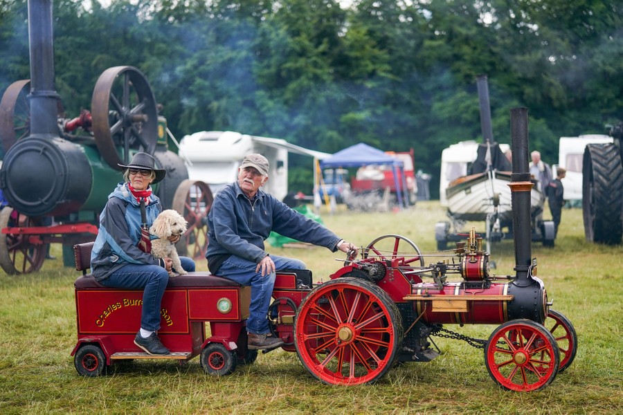 A couple and their dog drive a miniature steam engine on a grassy field.