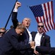 Former President Donald Trump is surrounded by Secret Service agents, backdropped by a large American flag, after an attempted assassination. Trump, with some visible blood on his face, raises his fist high.