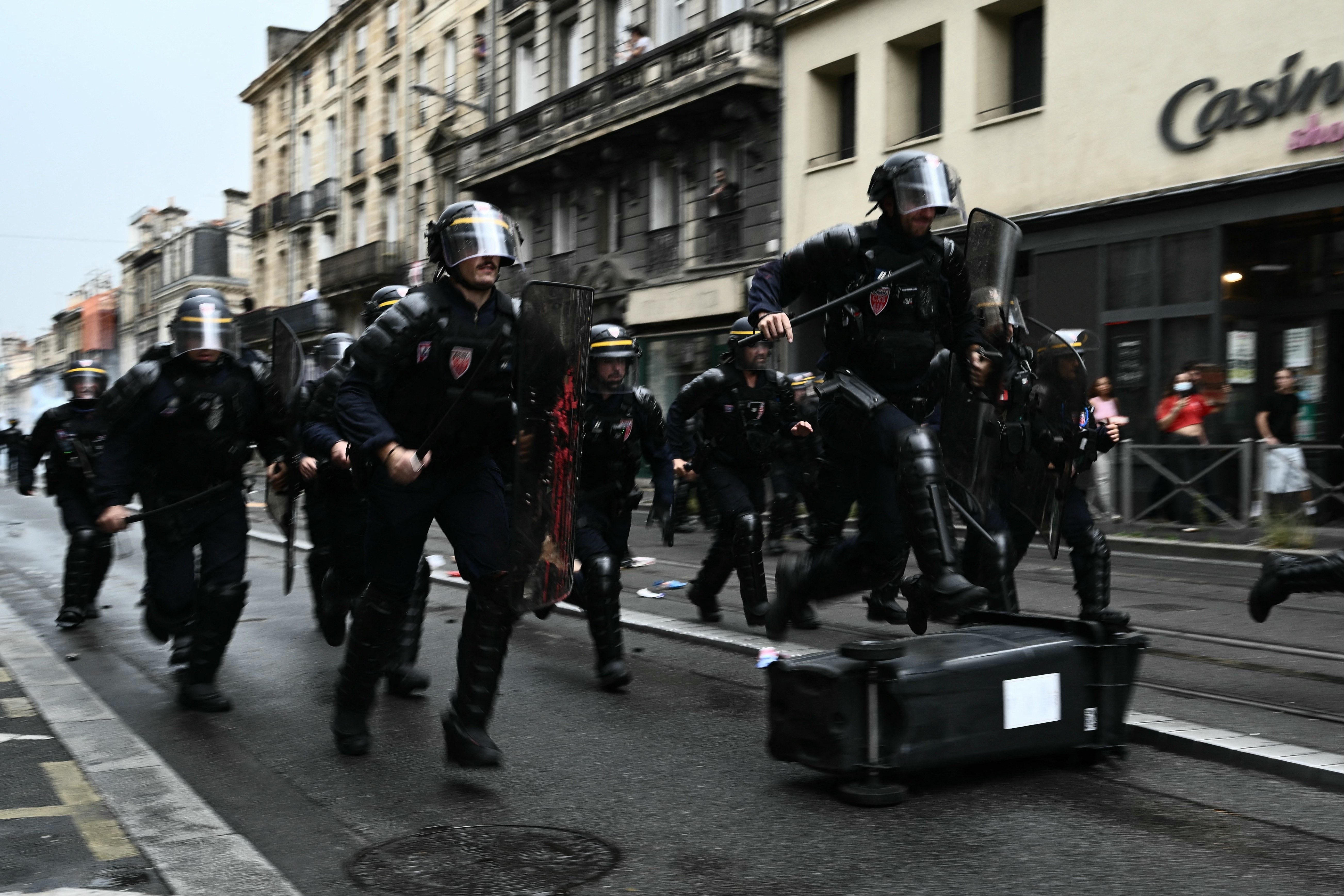 A group of riot police in full gear run down a street. One hurdles a toppled garbage bin.