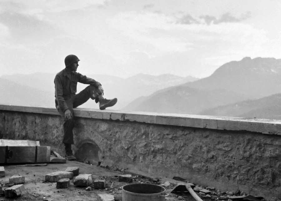 An American soldier rests on the edge of a balcony at the ruins of Berchtesgaden, looking out over a mountain view.