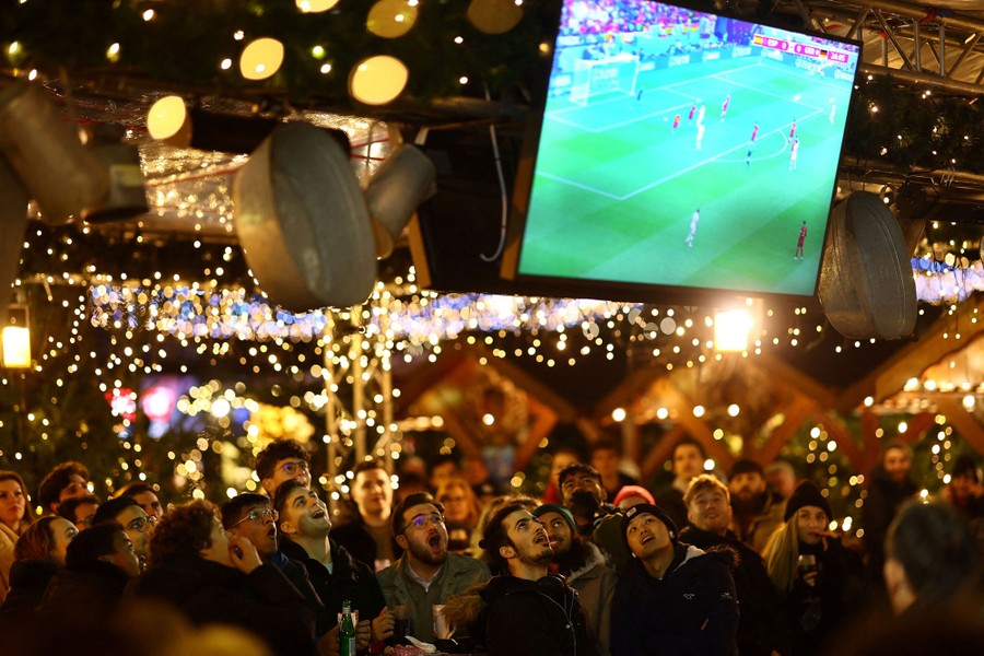 A group of people in a Christmas market look up at screens showing a soccer match.