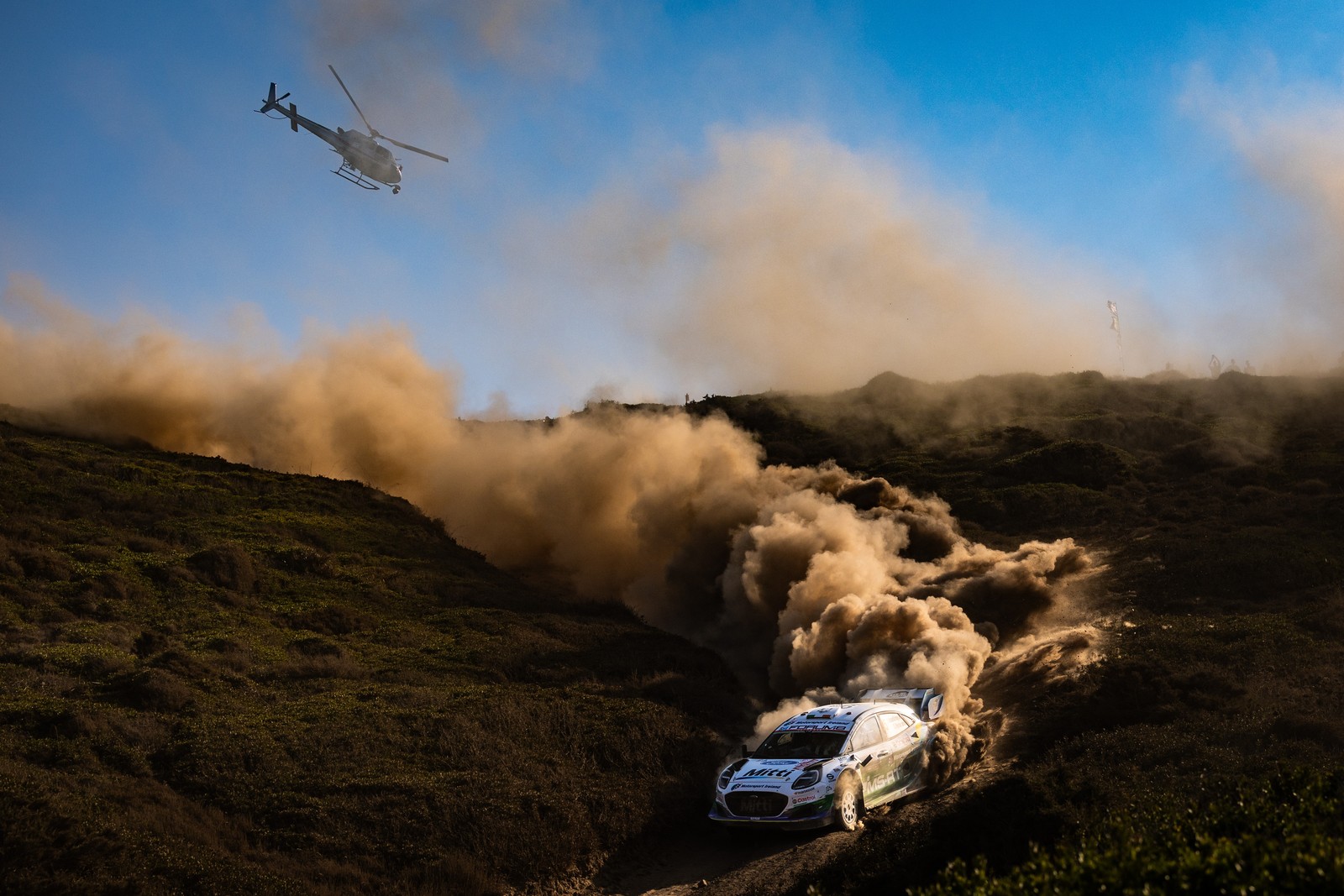 A helicopter chases a rally race car as it kicks up dust on a dirt road.