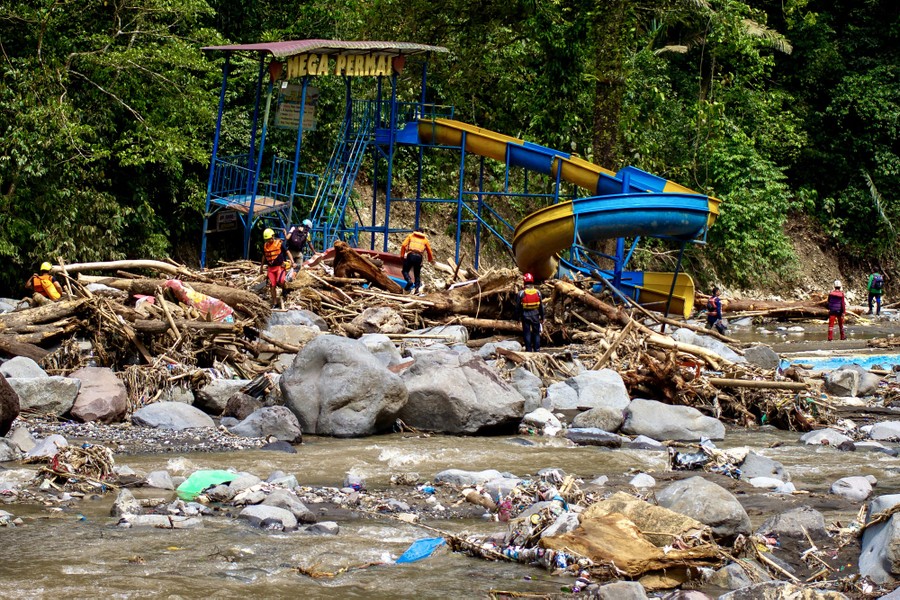 People search amid flood debris and the remains of a damaged amusement park ride along a river.