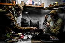 Two uniformed people sit at a table in a dimly lit room, handling wires and electronic equipment.