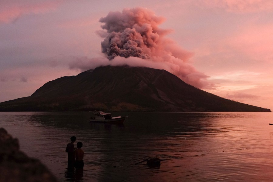 Two people stand on a shoreline, looking across water toward a volcano with clouds of ash erupting out of its peak.