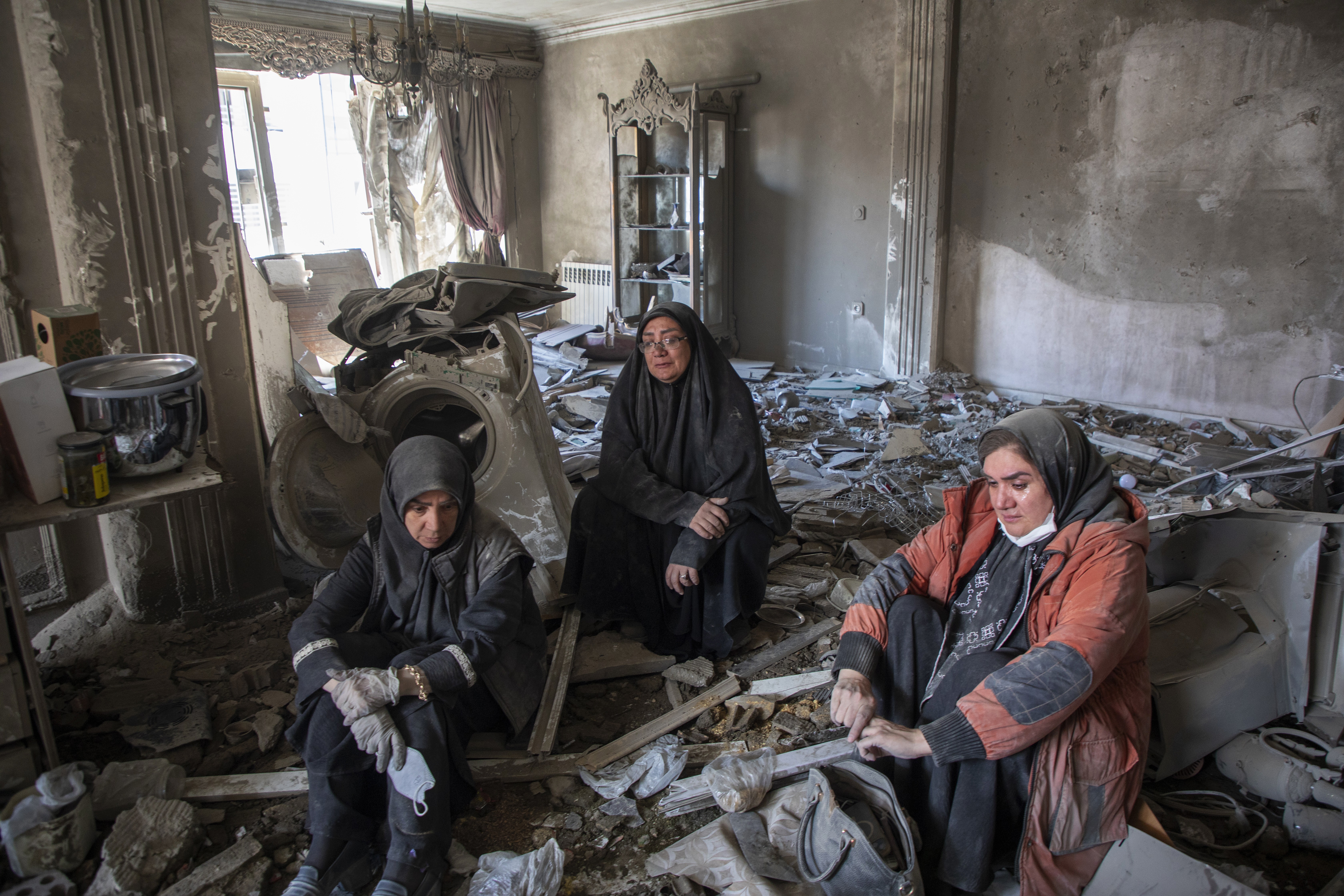 Three people sit inside a heavily-damaged room, among dust and debris, after an airstrike.