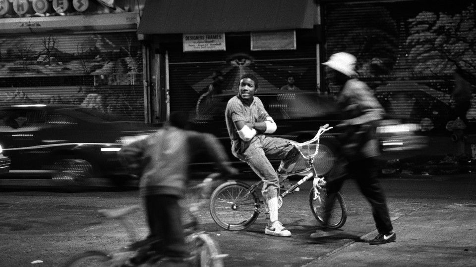 A Black teenager, in focus, sits on a bike with his arms folded, as people and cars around him are blurred.