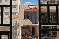 Photograph taken behind iron window grills of a destroyed building with an Iranian flag hanging from its side