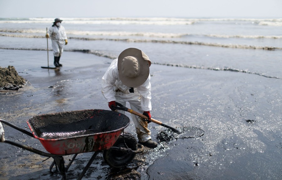 A person shovels a small amount of spilled oil from a contaminated beach into a wheelbarrow.